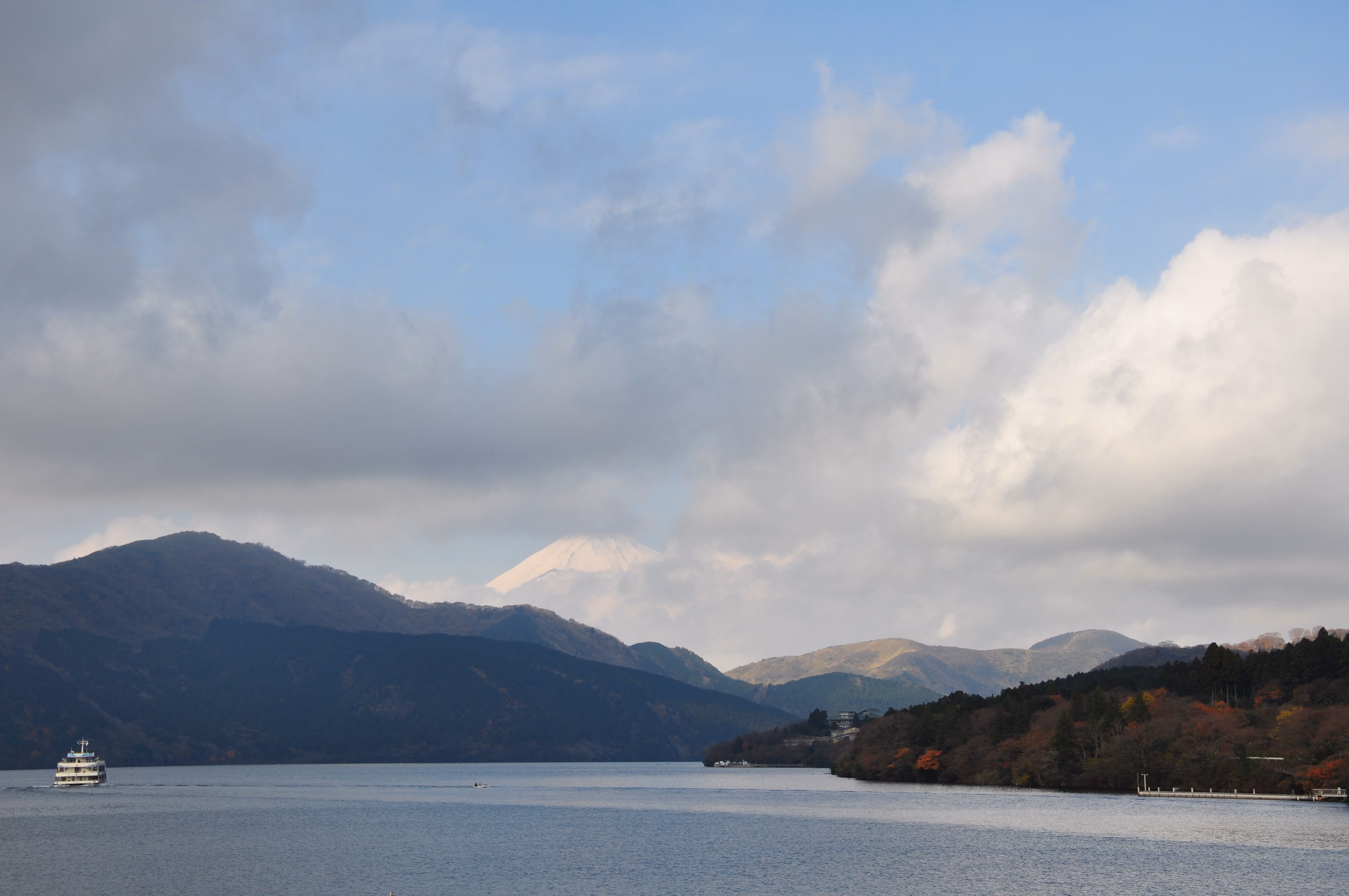 Mount Fuji's snow-capped peak rising above clouds, seen from Lake Ashi with a ferry on the water