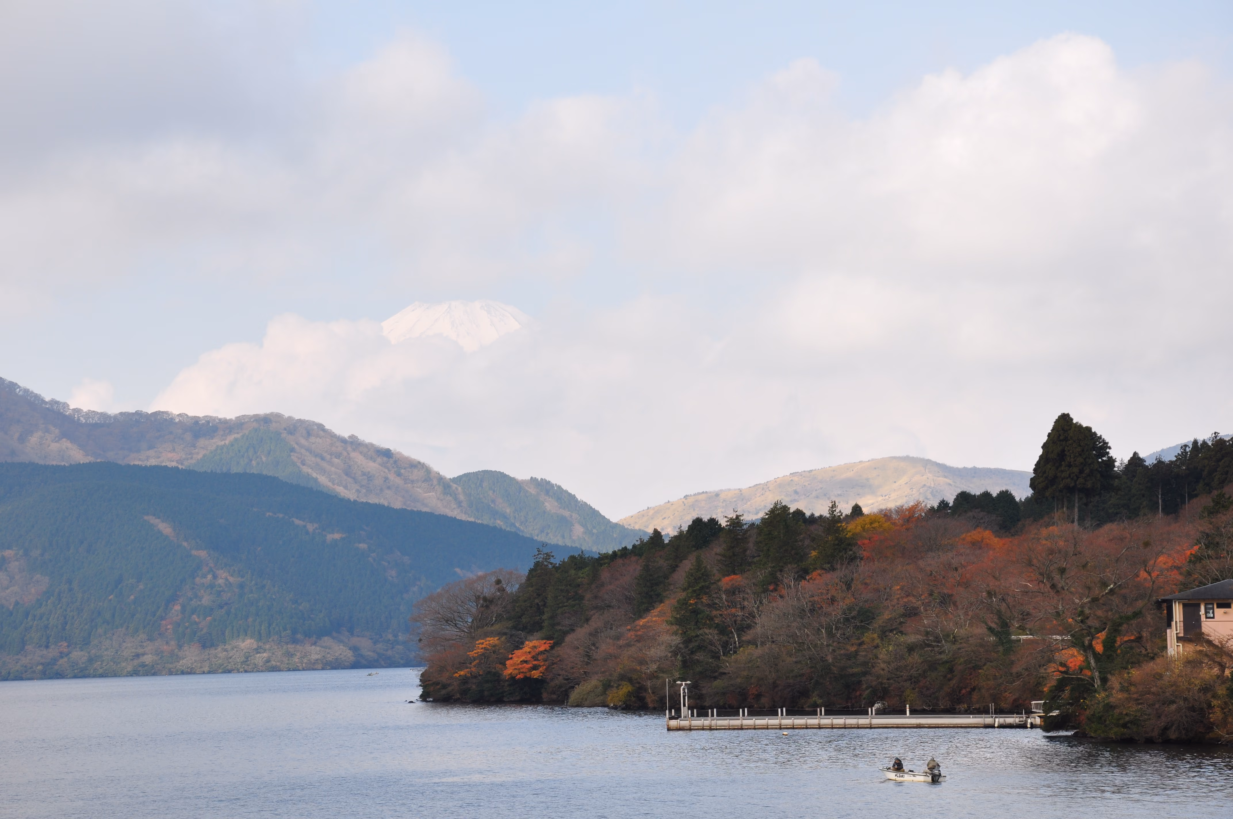 Mount Fuji's snow-capped peak above clouds, seen past a lone fishing boat on Lake Ashi with autumn foliage in the foreground