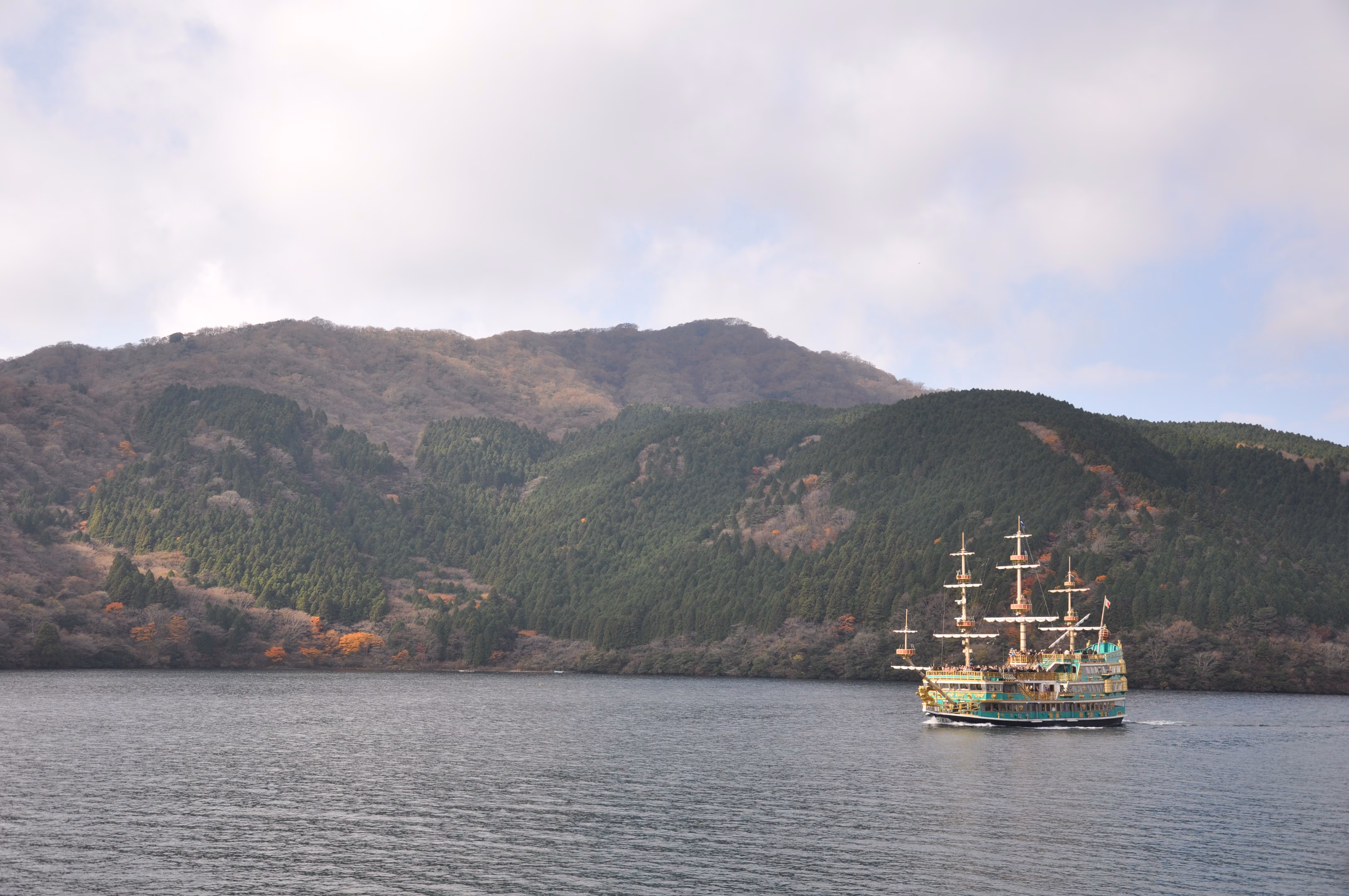 A green pirate-ship-styled tour boat crossing Lake Ashi with autumn hillside behind it