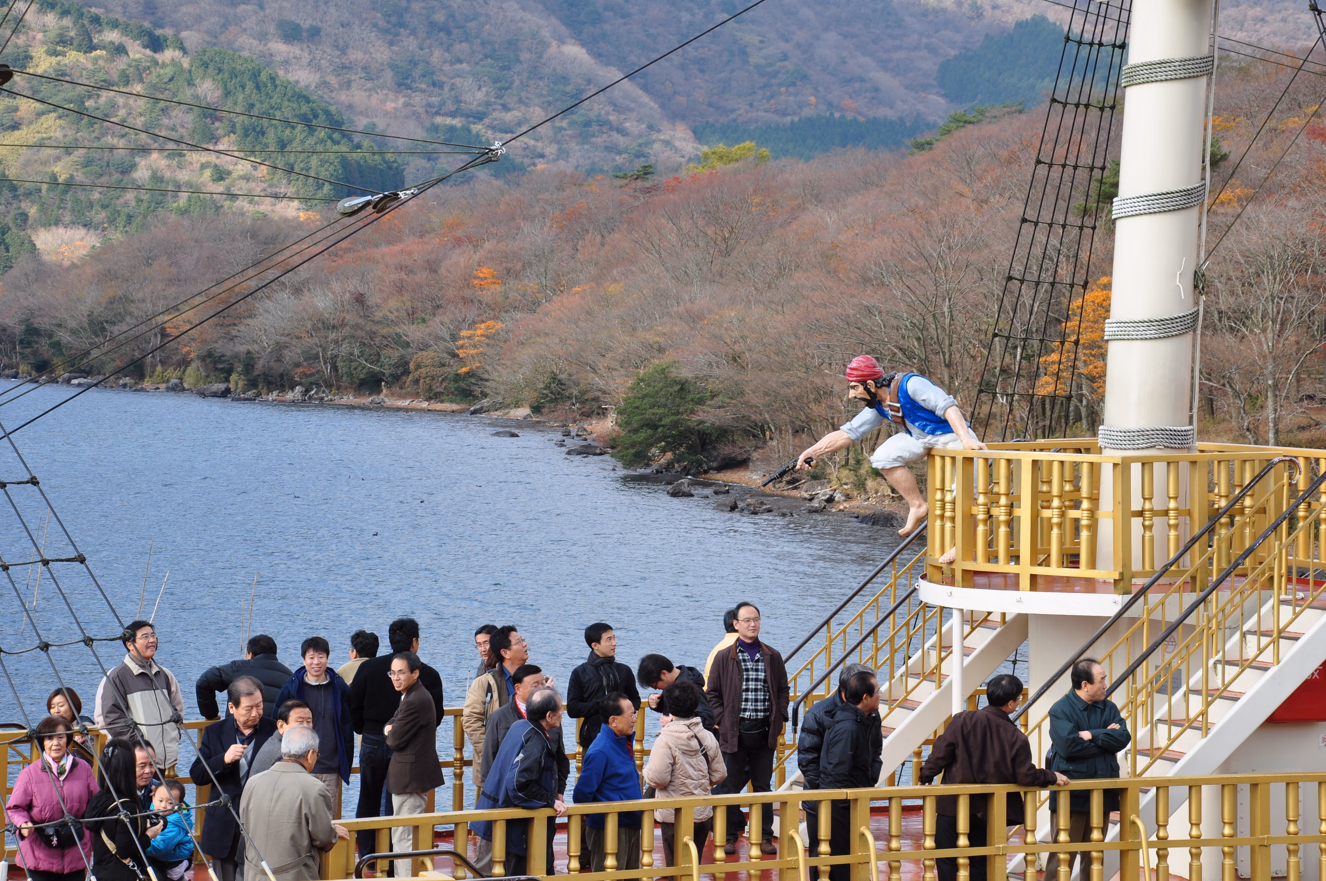 A pirate mannequin figure leaping over the rail of a tour boat, with passengers standing on deck below and Lake Ashi behind