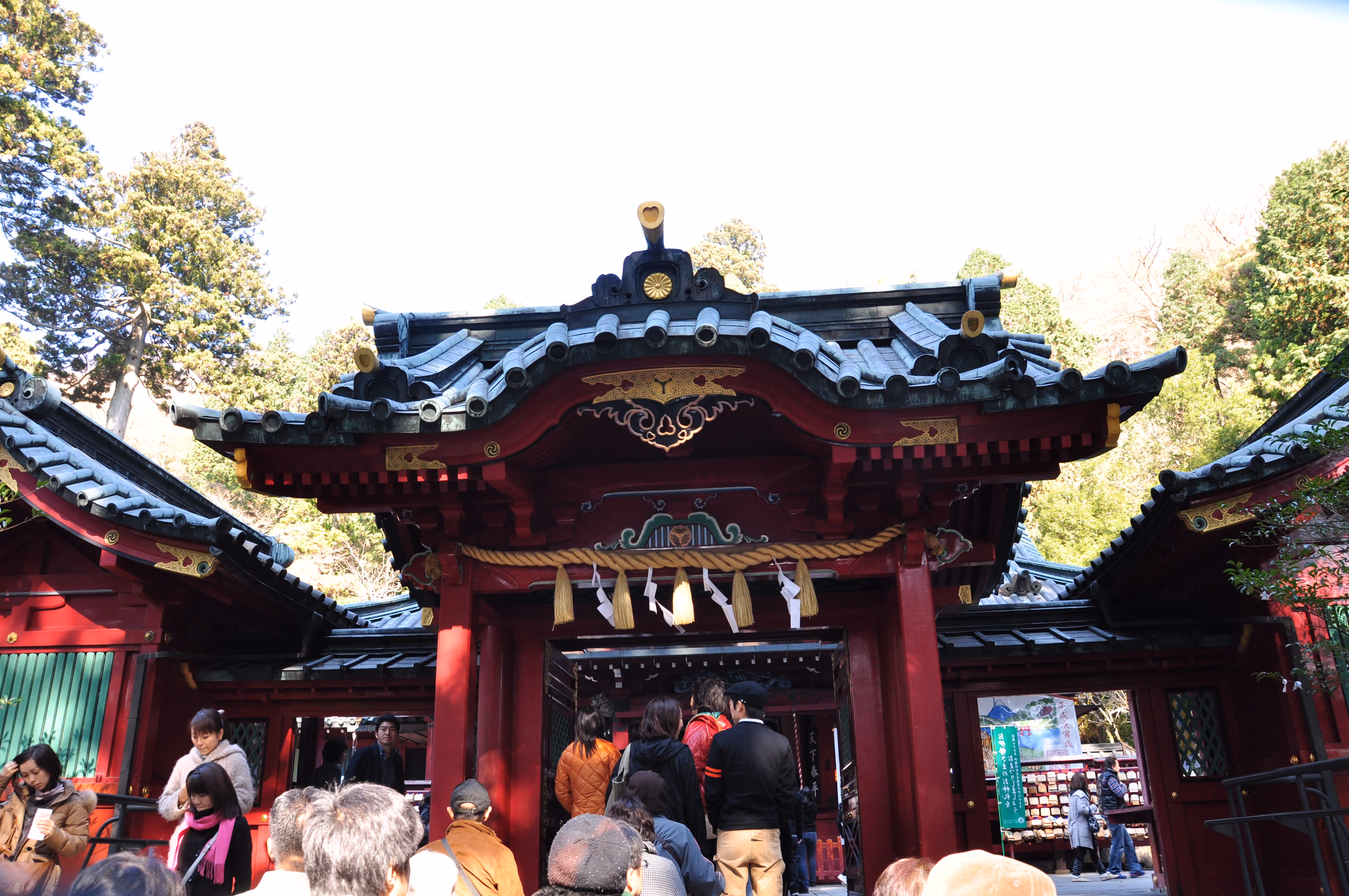 A red lacquered shrine gate with a sacred rope hung across it, with visitors queuing underneath