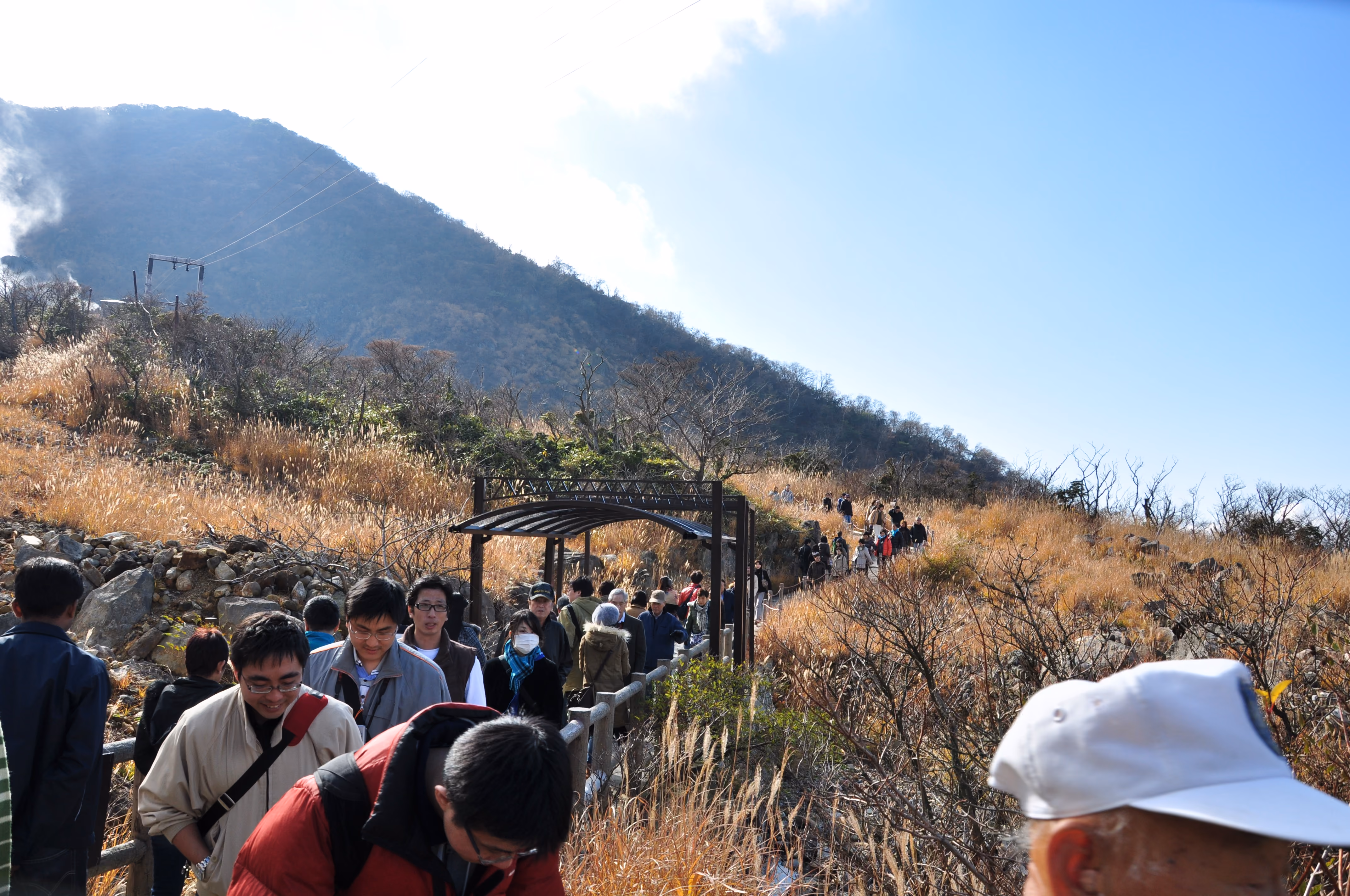 A crowd of visitors walking single file along a boardwalk path toward steam vents at Owakudani