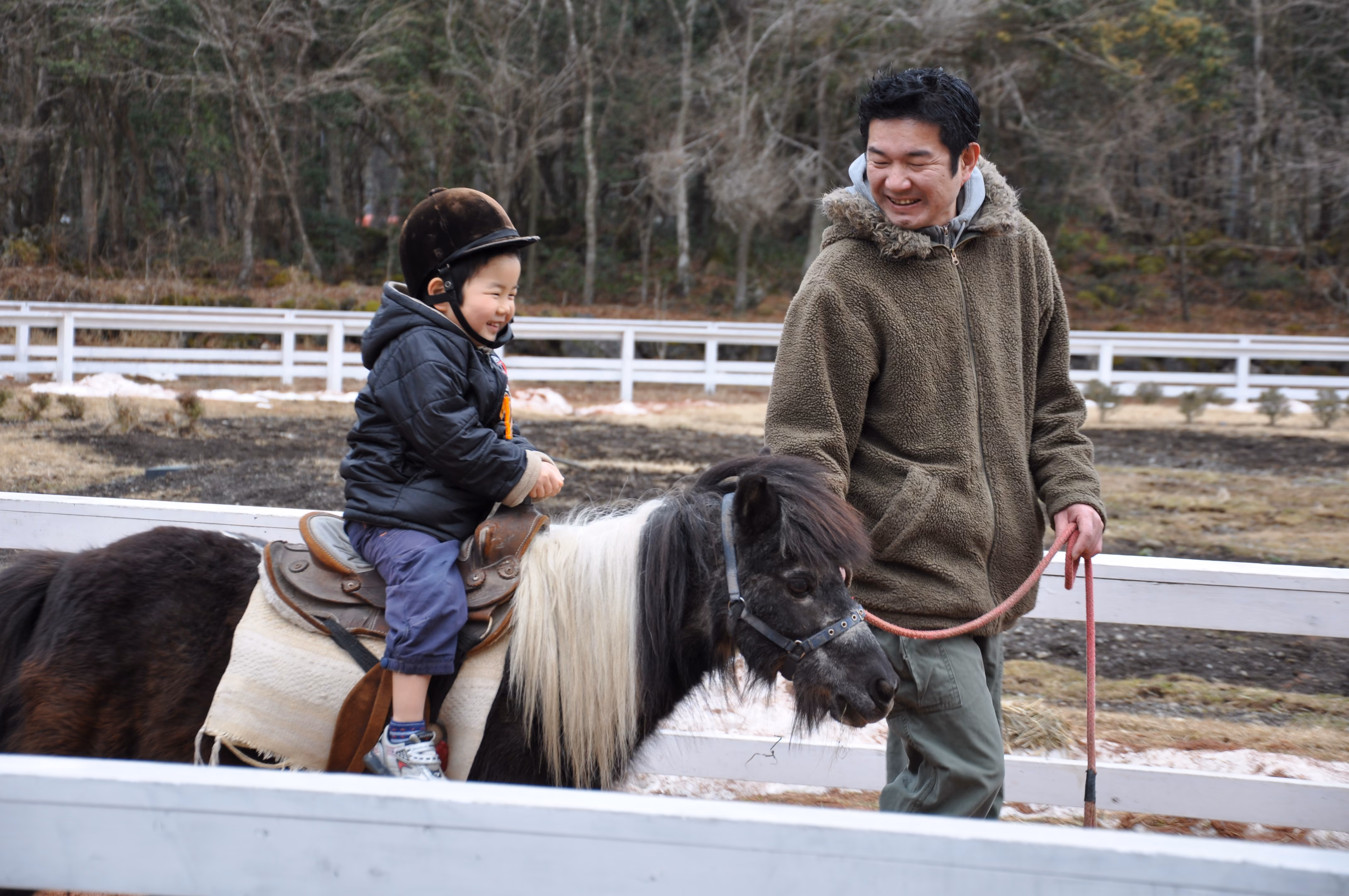 A child riding a pony led by an adult along a white fence