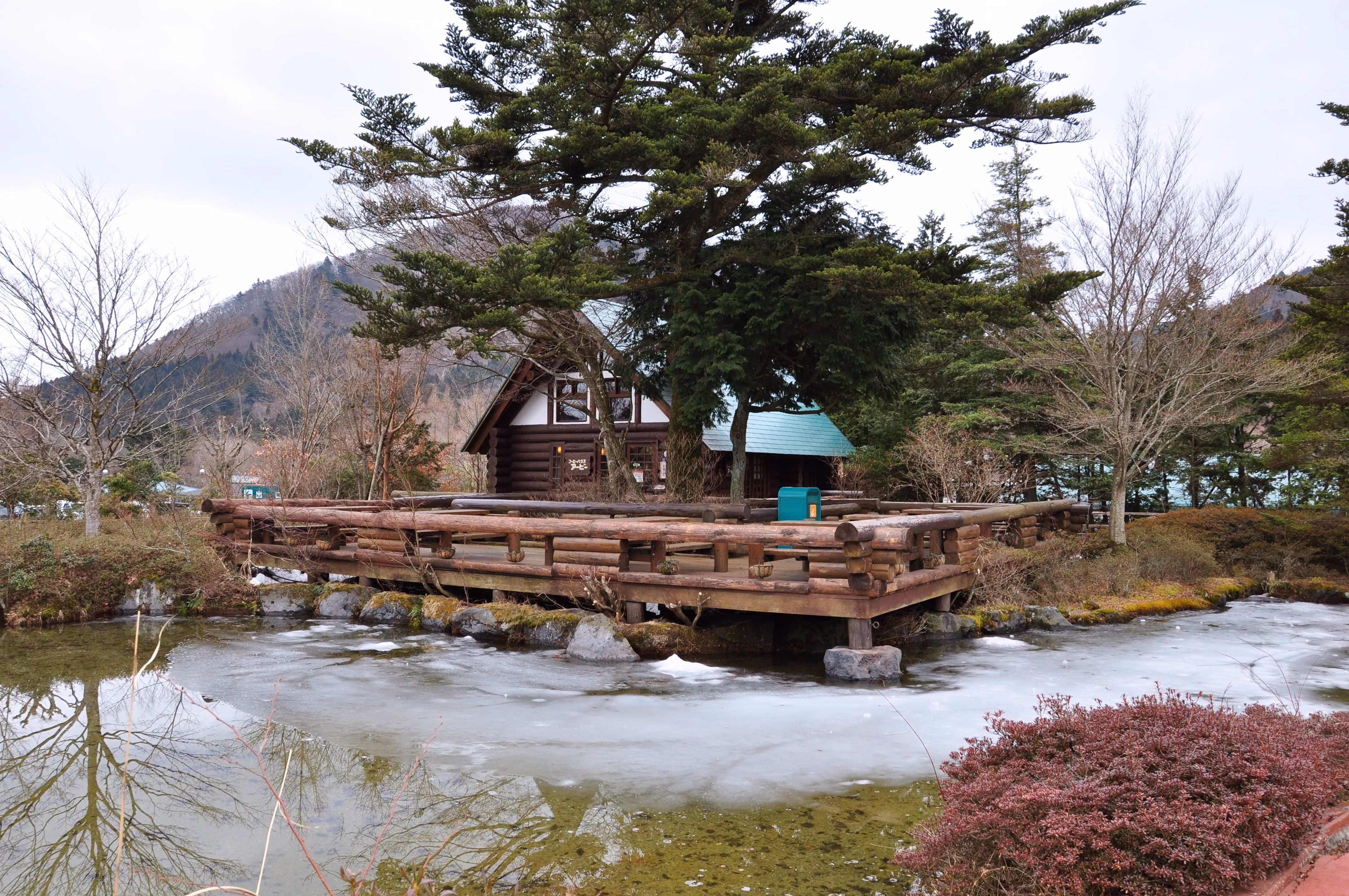 A log cabin lodge with a green roof on a small pond partially covered in ice, mountains in the background