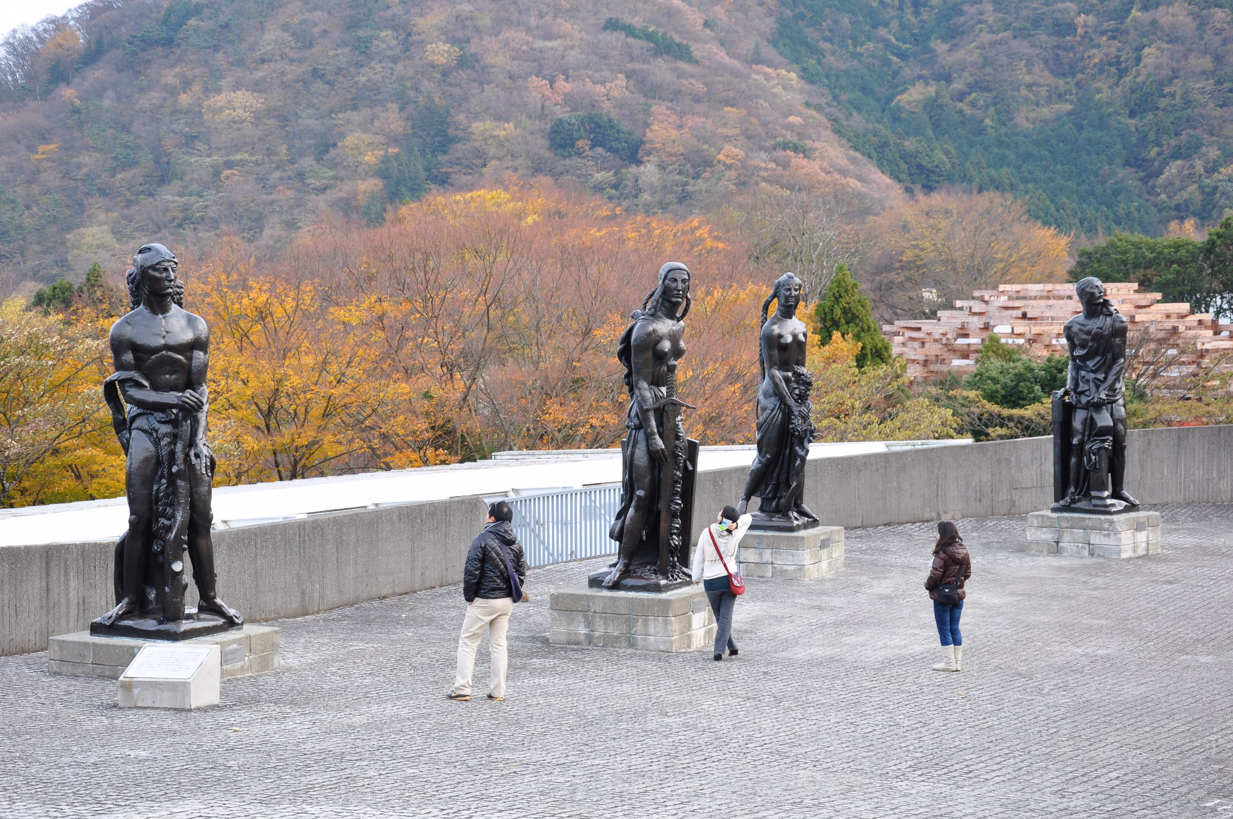 A row of large bronze figurative sculptures on an outdoor museum terrace, with an autumn hillside behind them