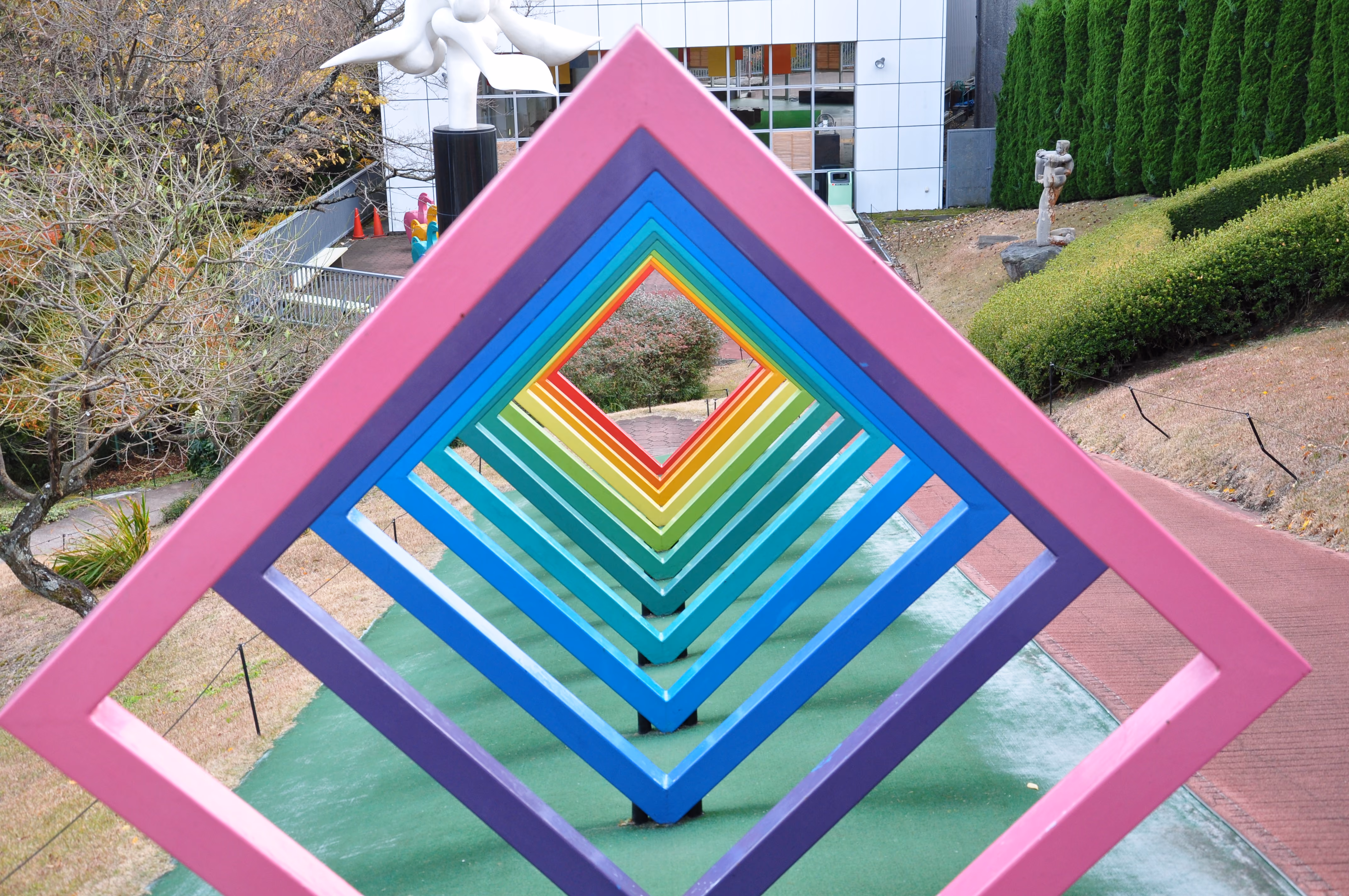 A walk-through sculpture of nested rainbow-colored diamond frames forming a tunnel, in a museum garden