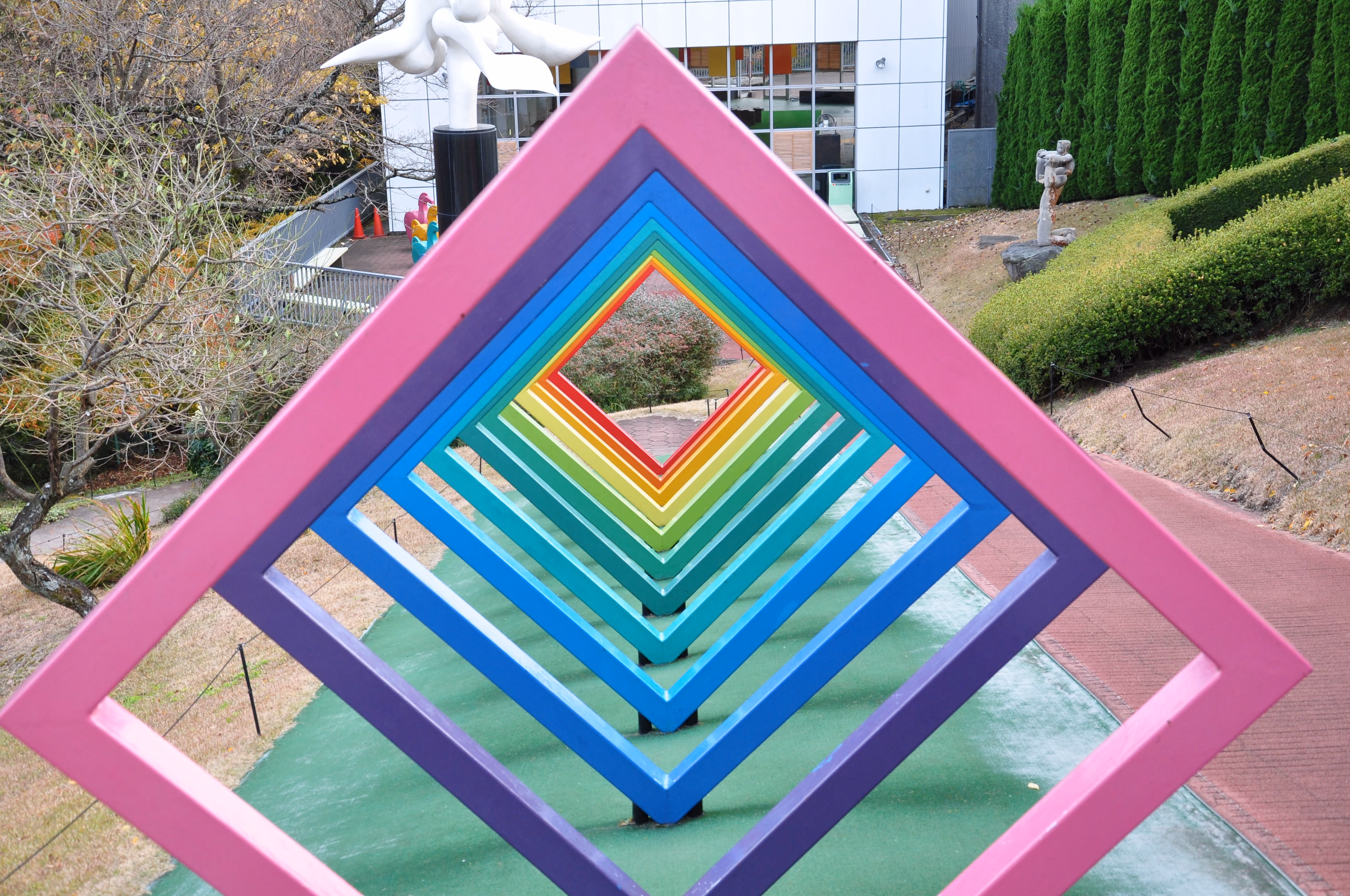 A walk-through sculpture of nested rainbow-colored diamond frames forming a tunnel, in a museum garden