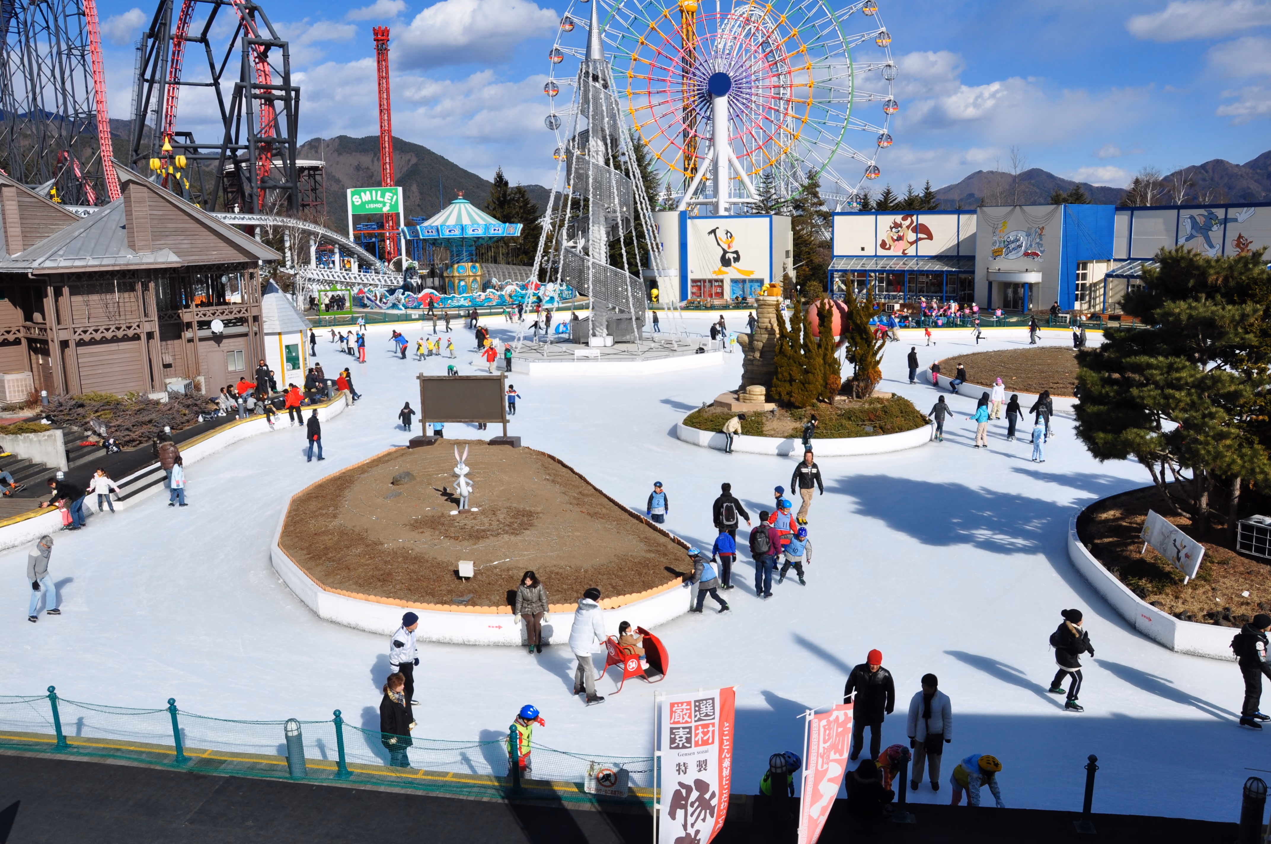 Wide view of an outdoor ice rink with cartoon murals on a nearby building and a ferris wheel in the background