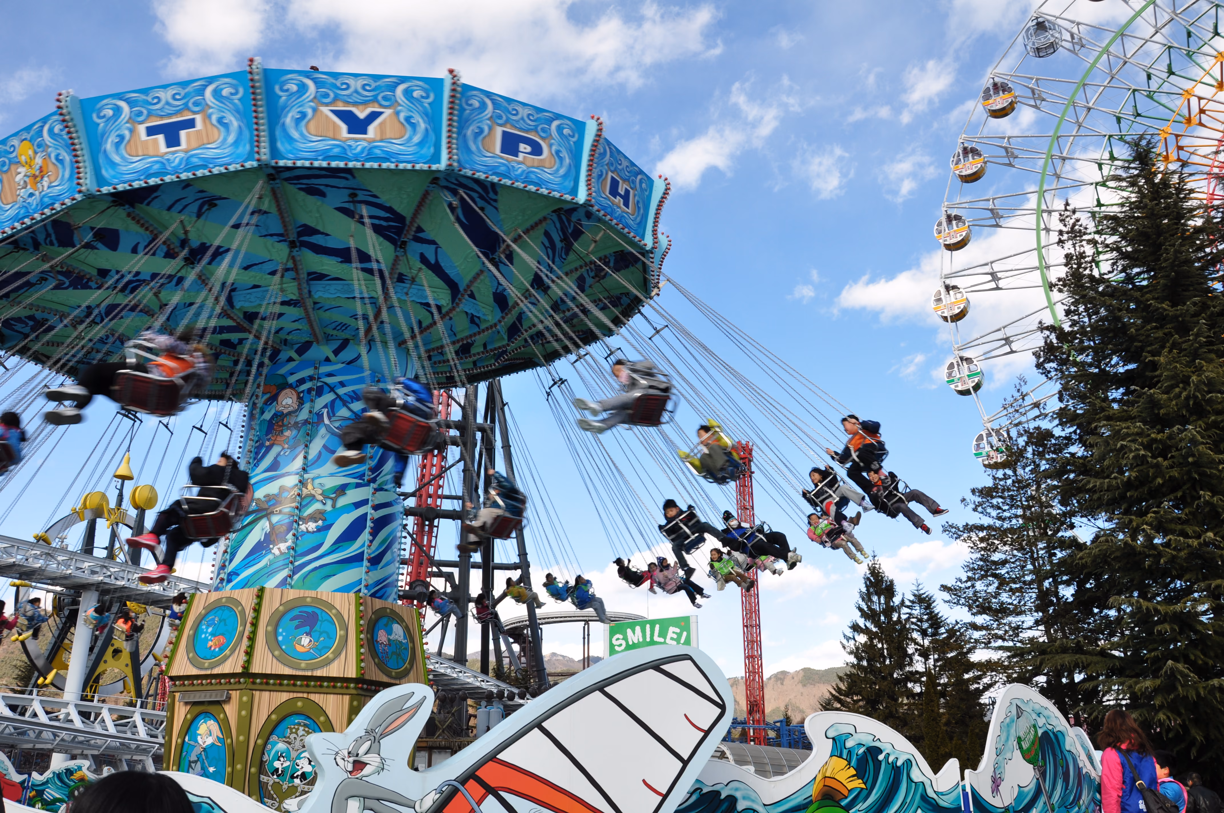 A swing carousel ride called Typhoon with riders swinging out against a blue sky, a ferris wheel behind it