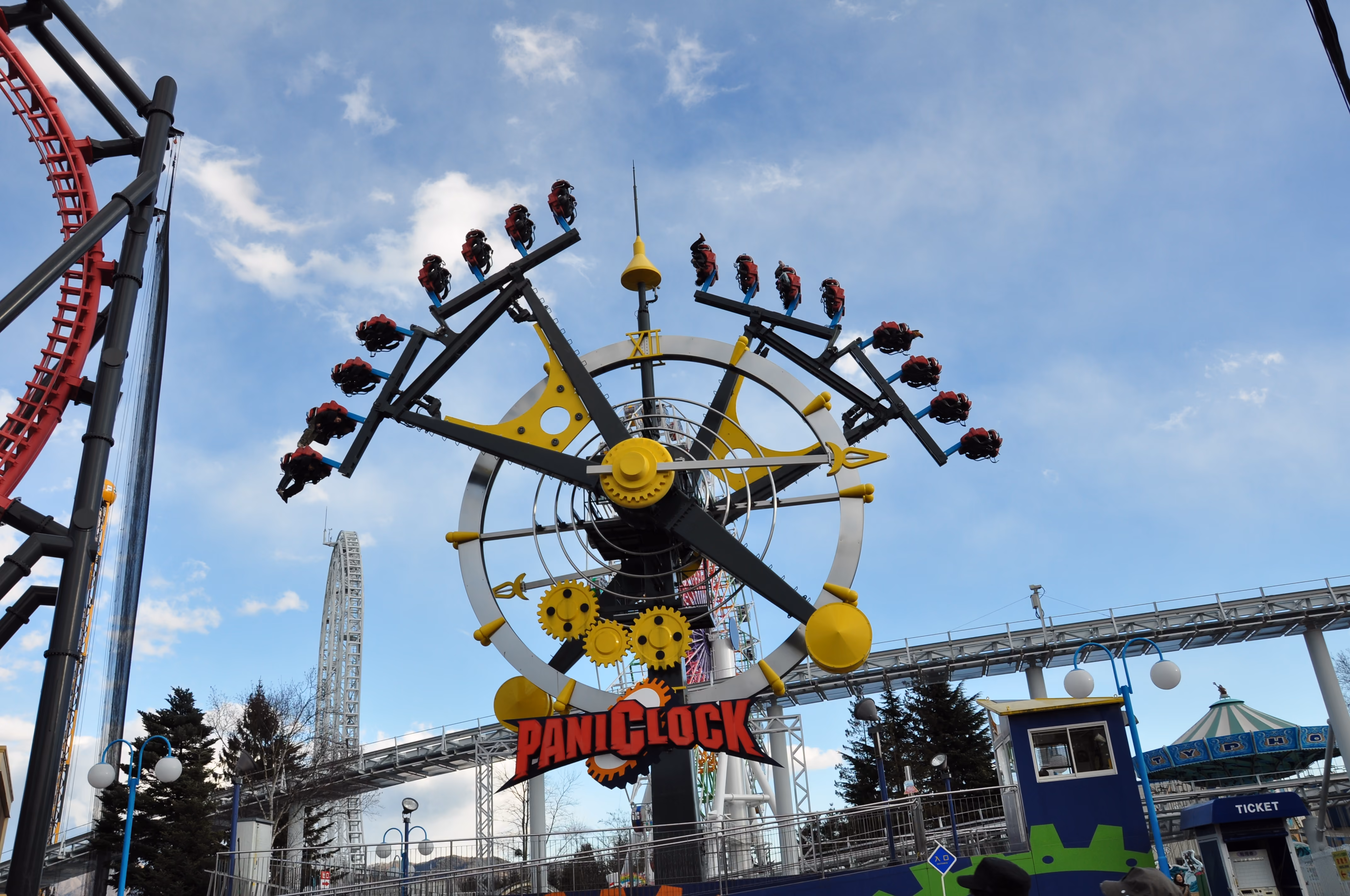 A clock-themed pendulum ride called PaniClock with riders suspended from clock hands against a blue sky