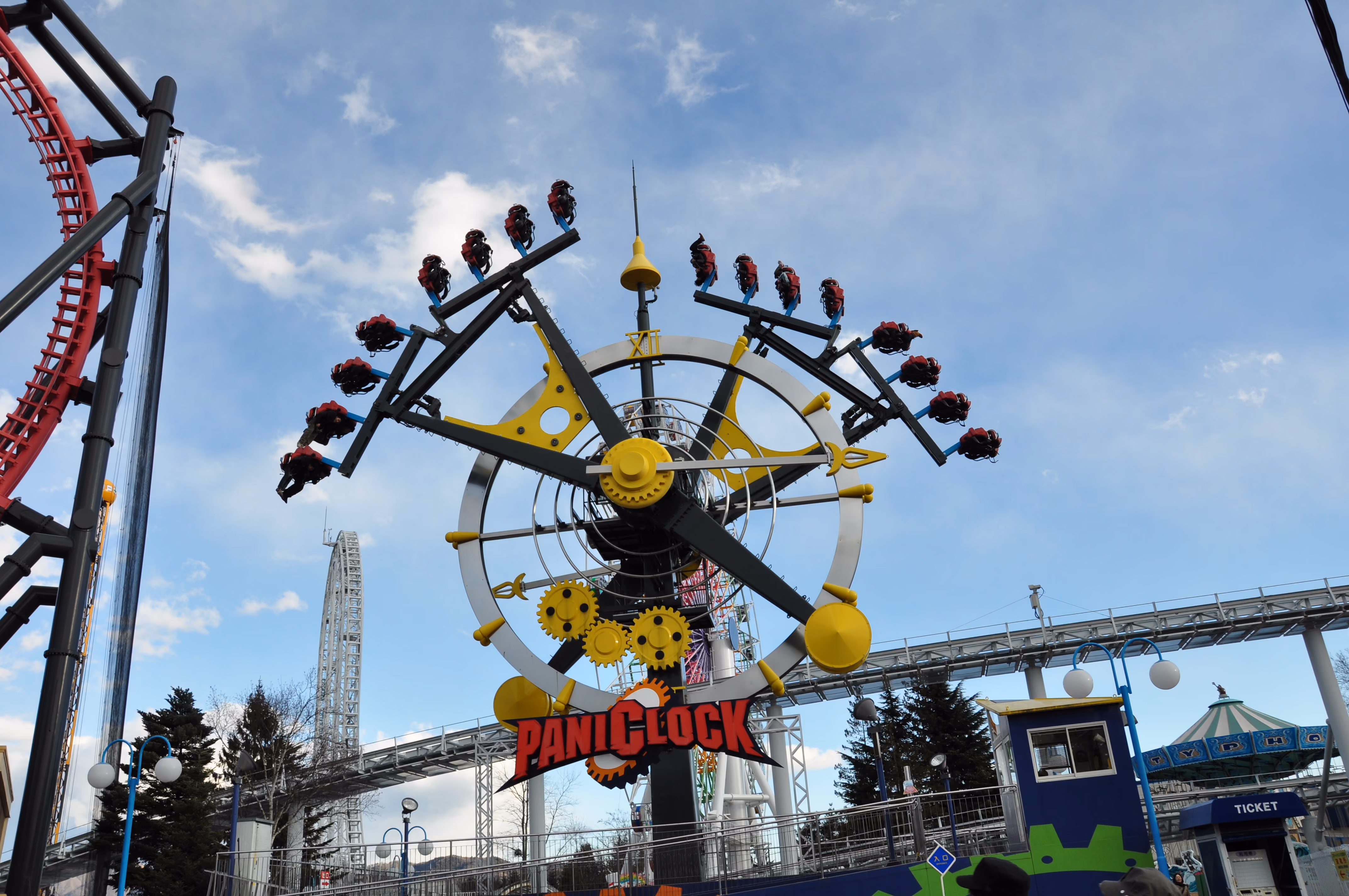 A clock-themed pendulum ride called PaniClock with riders suspended from clock hands against a blue sky