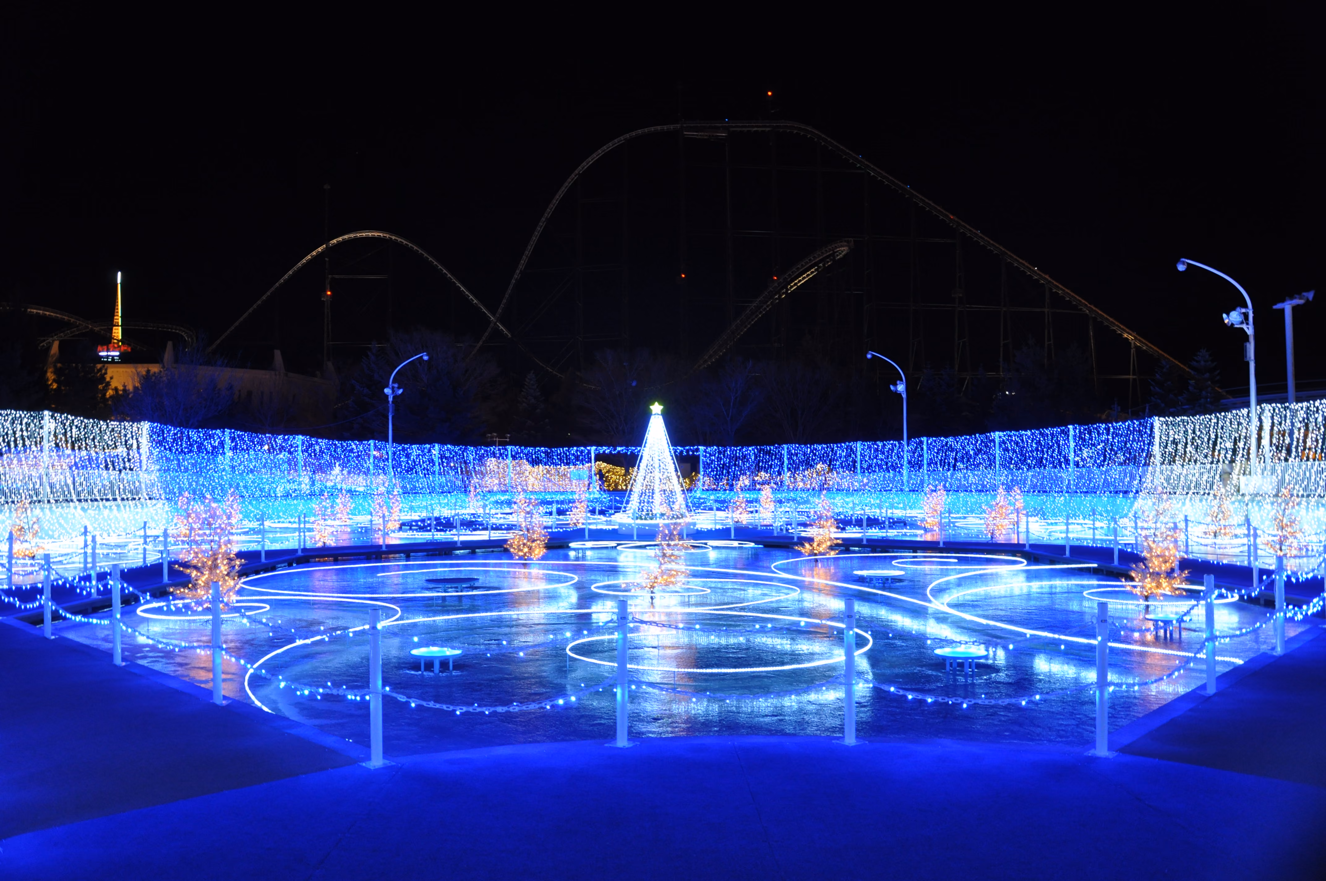 The outdoor ice rink at Fuji-Q Highland illuminated in blue light at night, with a lit tree in the center and a roller coaster silhouette behind it